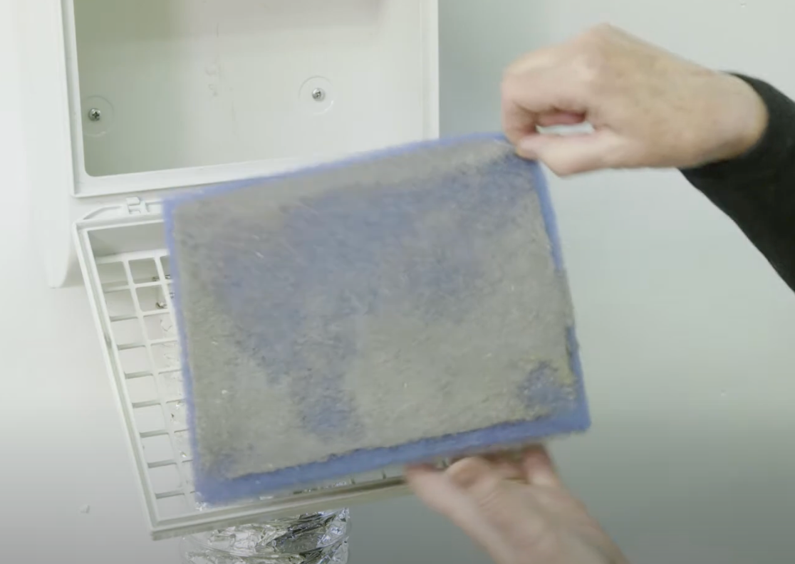Close-up of a person holding a used, lint-covered filter removed from a BetterVent indoor dryer vent. The vent housing is open in the background, showing the internal grate and duct connection.